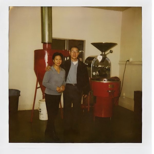 nwdn_file_temp_1608669478489 2 Mother and father standing in front of a red coffee roasting machine.